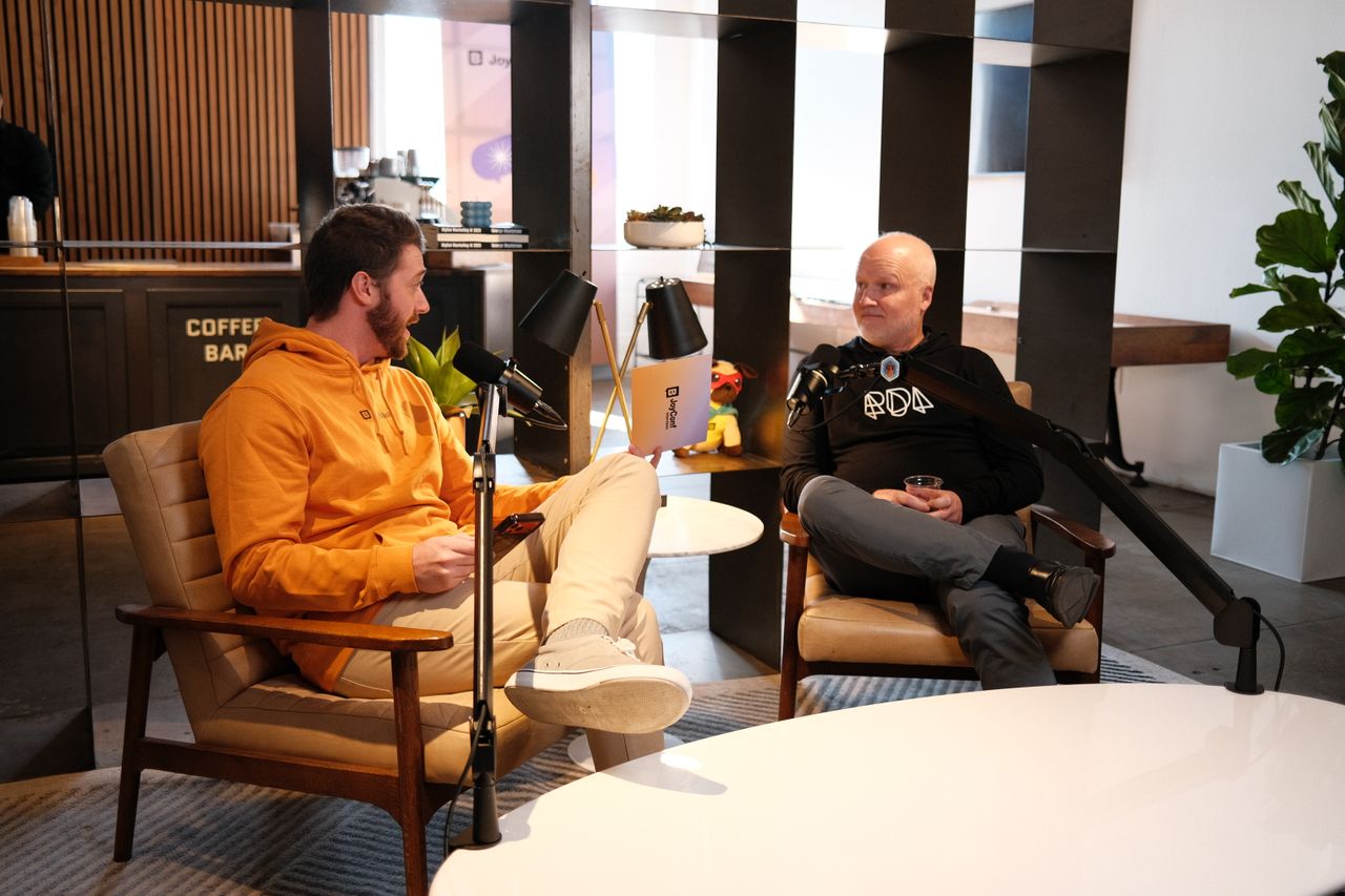 “Two men sit in armchairs recording a podcast-style conversation in a bright, modern café setting. The man on the left wears an orange hoodie and light pants, holding a phone and speaking into a microphone. The man on the right wears a black hoodie with a geometric logo and gray pants, holding a drink while responding into his microphone. A small round table sits between them, and shelves, plants, and a coffee bar are visible in the background.