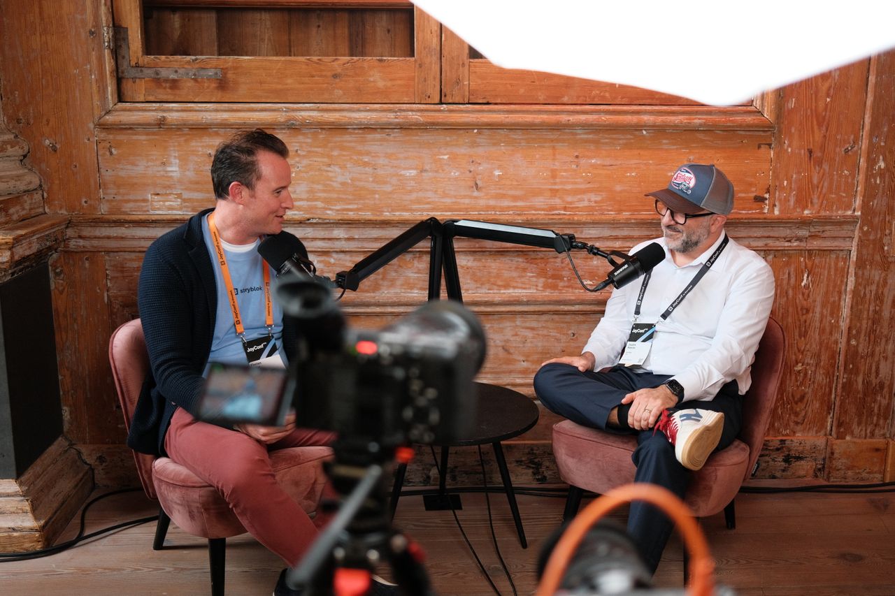 Two speakers recording a podcast-style interview during JoyConf, sitting across from each other in armchairs with microphones in a cozy wooden room.