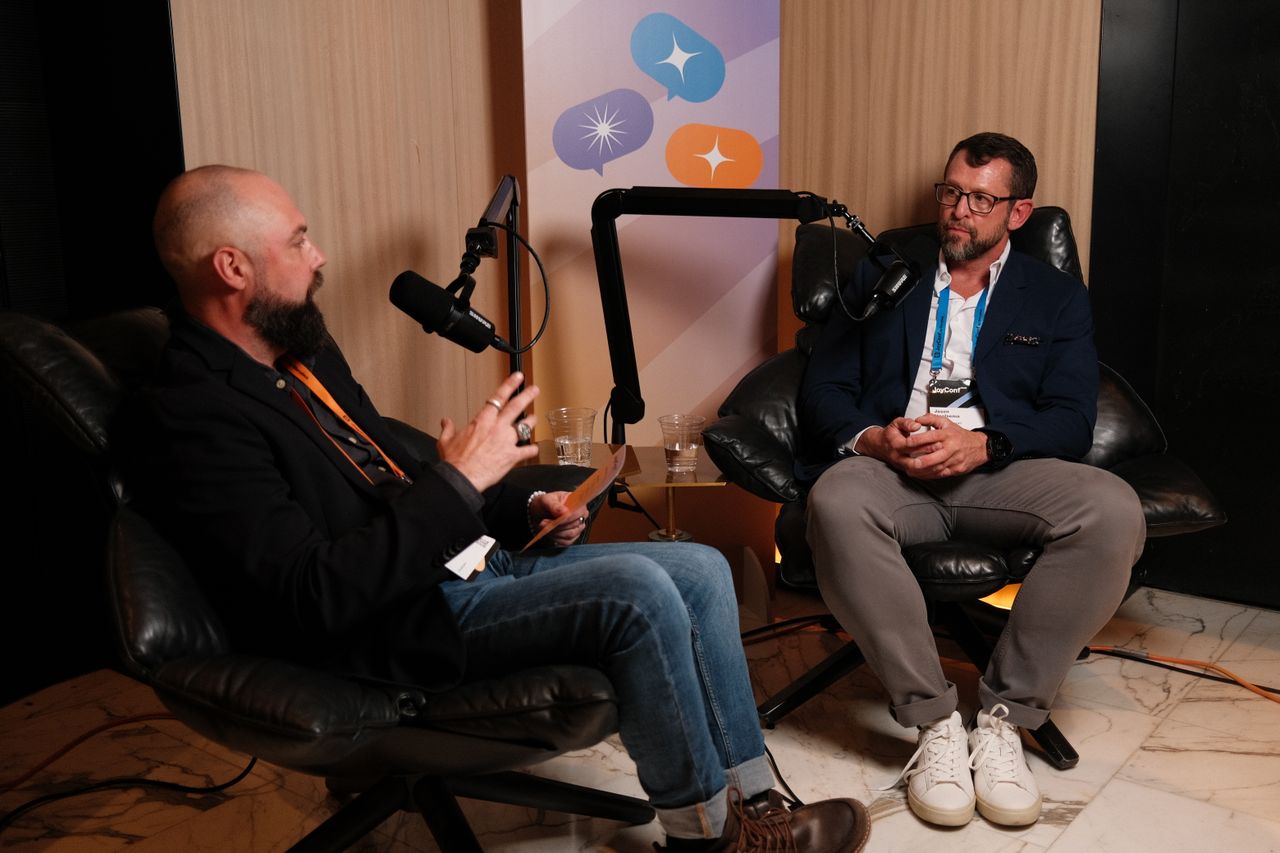 Two men sit in leather chairs recording a podcast-style conversation in a studio setup. The man on the left, bald with a beard, wears a black blazer, jeans, and brown boots, holding a notepad and speaking into a microphone. The man on the right wears glasses, a blue blazer, a white shirt, and gray pants, listening and preparing to respond. Multiple cameras and audio equipment are set up in the foreground, and a banner with colorful speech bubble icons hangs behind them.