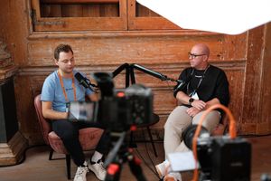 Two speakers recording a podcast-style interview during JoyConf, sitting across from each other in armchairs with microphones in a cozy wooden room.