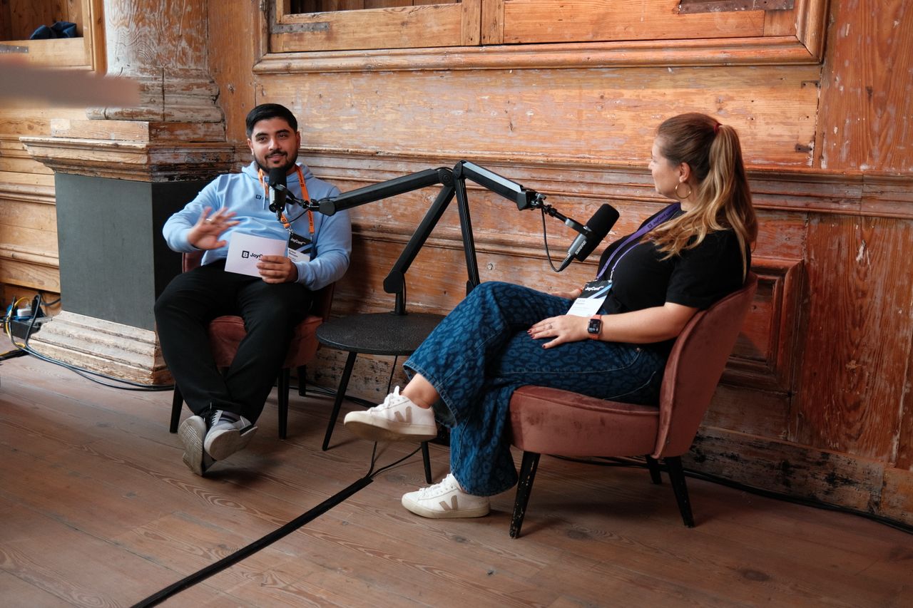 Two speakers recording a podcast-style interview during JoyConf, sitting across from each other in armchairs with microphones in a cozy wooden room.