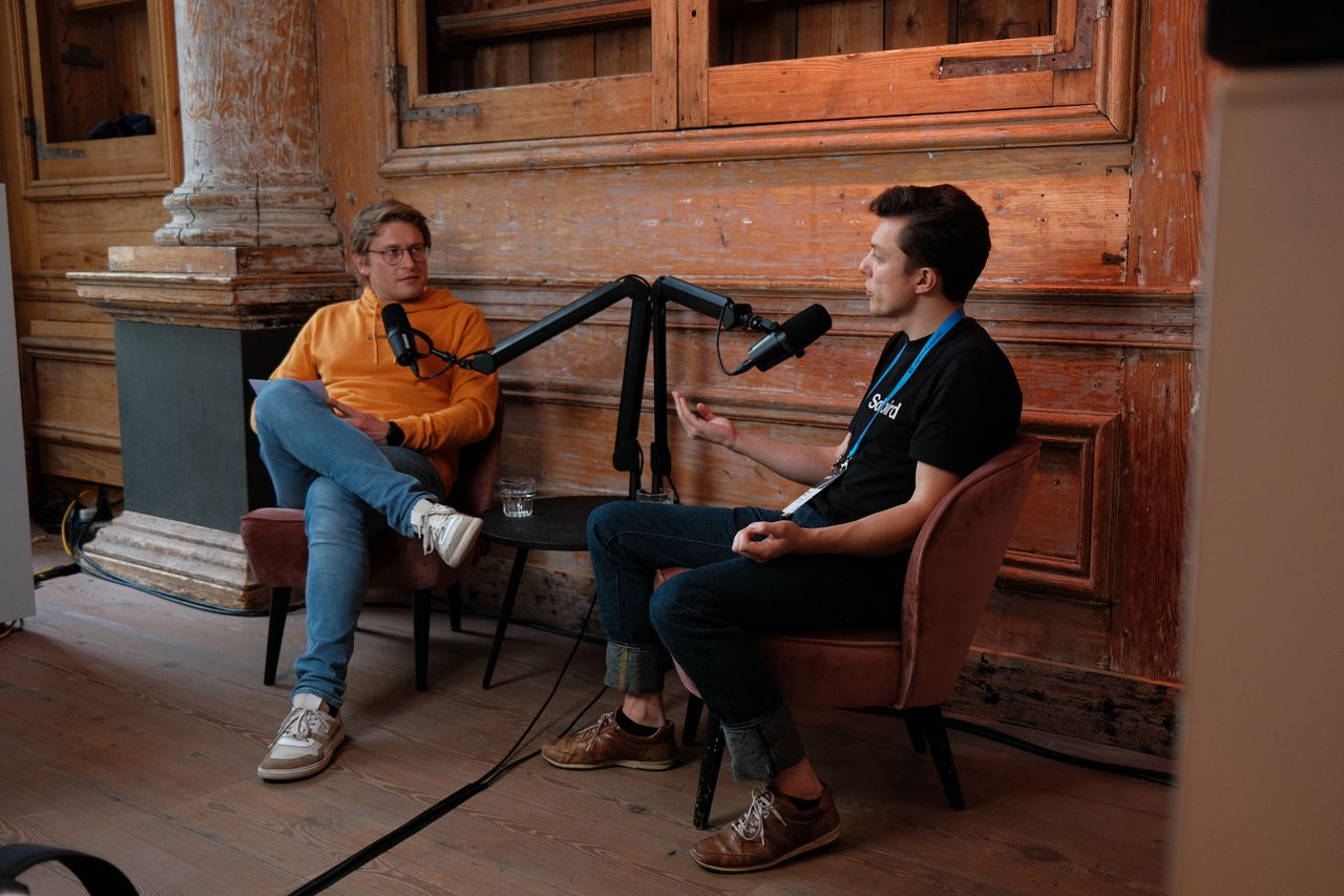 Two speakers recording a podcast-style interview during JoyConf, sitting across from each other in armchairs with microphones in a cozy wooden room.