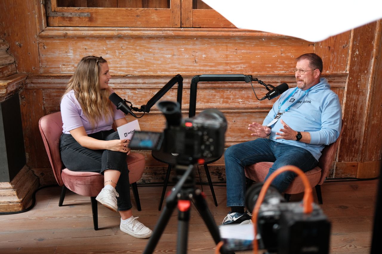Two speakers recording a podcast-style interview during JoyConf, sitting across from each other in armchairs with microphones in a cozy wooden room.
