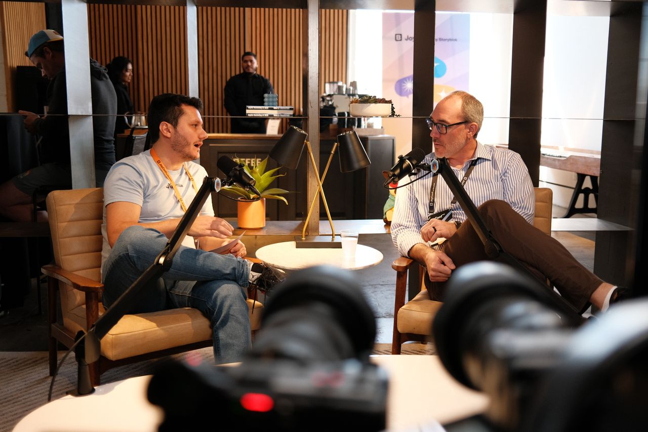 Two men sit in armchairs recording a podcast-style conversation in a cozy café setting. The man on the left wears a light blue T-shirt, jeans, and an orange lanyard while speaking into a microphone and holding a notepad. The man on the right wears glasses, a striped shirt, and brown pants, listening and speaking into his own microphone. Camera equipment is visible in the foreground, and a coffee bar with people in the background adds to the casual atmosphere.