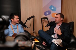 “Two men sit in leather chairs recording a podcast-style conversation with professional microphones. The man on the left wears a light blue hoodie and an orange lanyard, holding a tablet. The man on the right wears a blazer, button-up shirt, jeans, and sneakers, speaking while gesturing with one hand. A camera is visible in the foreground, and a banner with colorful speech bubble icons hangs behind them.
