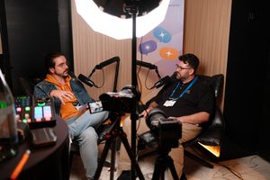 “Two men sit across from each other in leather chairs recording a podcast-style interview. The man on the left wears glasses, a yellow hoodie, a denim jacket, and light jeans, holding a laptop on his lap while speaking into a microphone. The man on the right wears glasses, a black polo shirt, and khaki pants, listening and preparing to respond into his microphone. Multiple cameras and audio equipment are set up in the foreground, and a banner with colorful speech bubble icons hangs behind them.