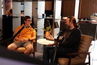 Two men sit in armchairs recording a podcast-style conversation. The man on the left wears glasses, an orange hoodie, and gray shorts, speaking into a microphone. The man on the right has long hair tied back, a beard, and wears a dark sweater and jeans, gesturing with his hands while talking. A small round table sits between them, and shelving, plants, and a coffee bar are visible in the background.