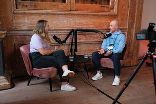 Two speakers recording a podcast-style interview during JoyConf, sitting across from each other in armchairs with microphones in a cozy wooden room.
