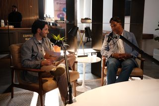 Two men sit in armchairs having a recorded conversation in a warmly lit café-style setting. The man on the left wears a light striped jacket and holds a JoyConf-branded notepad while speaking into a microphone. The man on the right wears a light blue blazer, patterned shirt, and jeans, responding into another microphone. A small round table with a cup of water sits between them, while shelves, plants, and lamps decorate the background.