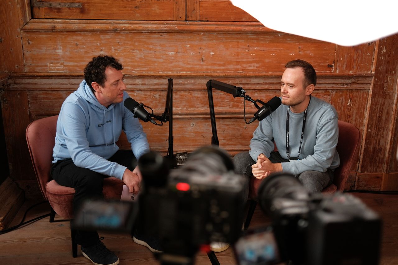 Two speakers recording a podcast-style interview during JoyConf, sitting across from each other in armchairs with microphones in a cozy wooden room.