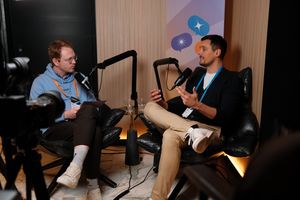 Two men sit across from each other in leather chairs recording a podcast-style conversation. The man on the left wears glasses, a light blue hoodie, dark pants, and white sneakers, holding a notepad with an orange lanyard around his neck. The man on the right gestures with his hands while speaking, wearing a dark cardigan, light pants, and white sneakers with a blue lanyard. Both speak into professional microphones, with a camera visible in the foreground and a banner with colorful speech bubble icons in the background.