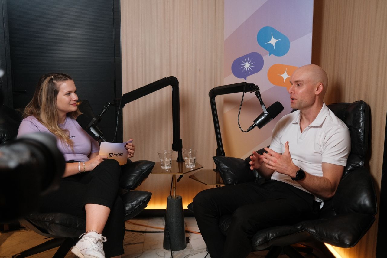 A woman and a man sit across from each other in leather chairs recording a podcast-style conversation. The woman on the left wears a lavender T-shirt, black pants, and white sneakers, holding a JoyConf-branded notepad and speaking into a microphone. The man on the right wears a white polo shirt and black pants, gesturing with his hands while talking into another microphone. A camera is visible in the foreground, and a banner with colorful speech bubble icons hangs behind them.