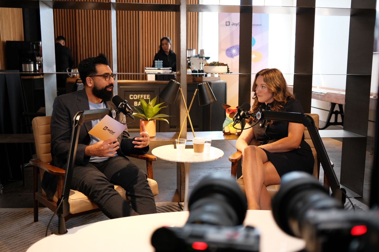 Two people sit in cushioned armchairs recording a podcast-style conversation in a cozy café setting. The man on the left, with dark hair, glasses, and a beard, wears a blazer and a light blue shirt while speaking into a microphone. The woman on the right has short brown hair and wears a denim jacket, green patterned shirt, and light pants, responding into her microphone. A small round table with cups of water sits between them, with shelves, plants, lamps, and a plush toy in the background.