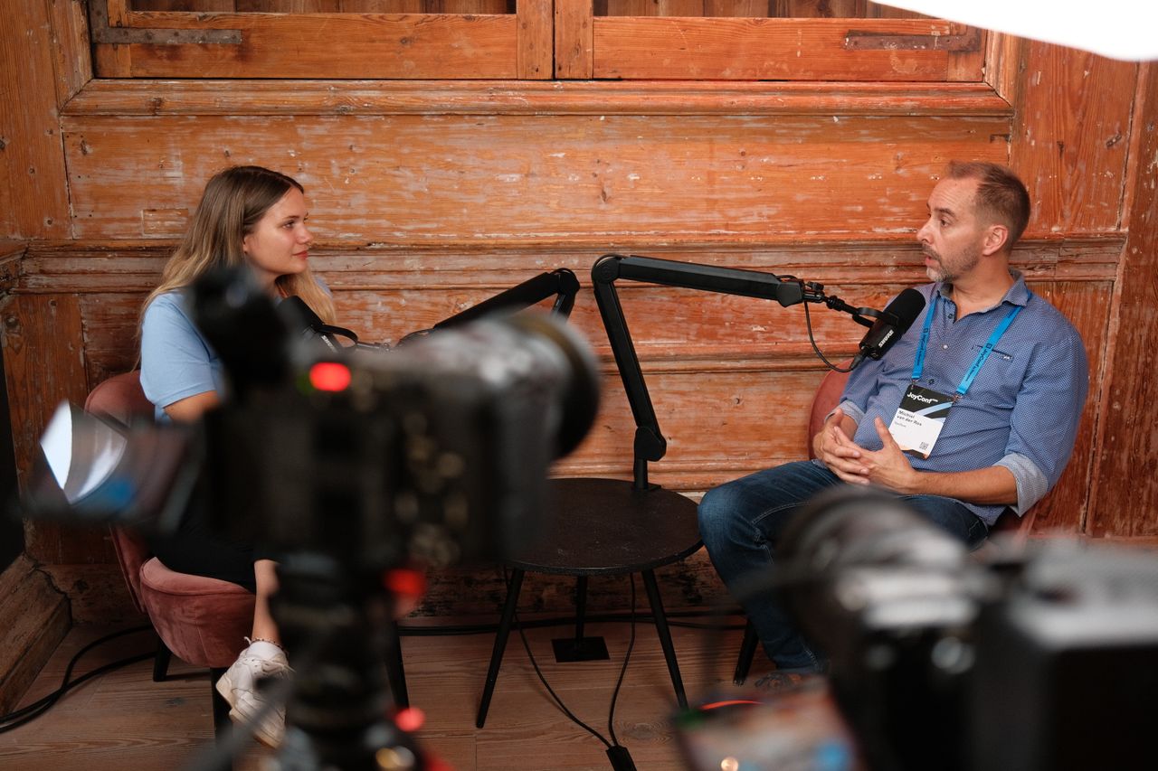 Two speakers recording a podcast-style interview during JoyConf, sitting across from each other in armchairs with microphones in a cozy wooden room.