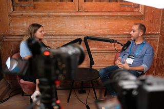 Two speakers recording a podcast-style interview during JoyConf, sitting across from each other in armchairs with microphones in a cozy wooden room.