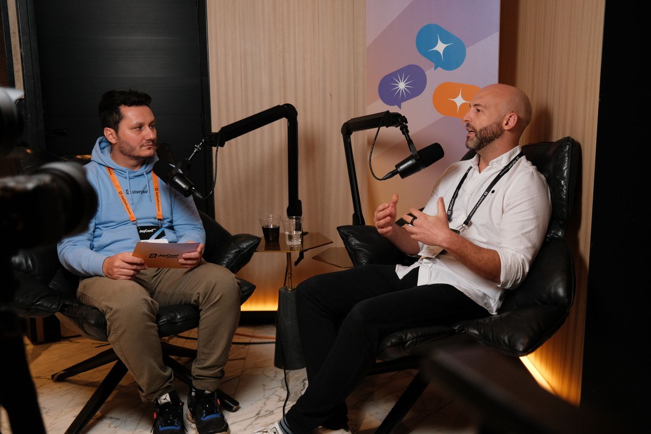 “Two men sit in leather chairs recording a podcast-style conversation. The man on the left wears a light blue hoodie, khaki pants, and an orange event lanyard, holding a notepad. The man on the right gestures while speaking into a microphone, wearing a white button-up shirt, black pants, and a black lanyard. A camera is visible in the foreground, and a banner with colorful speech bubble icons hangs behind them.