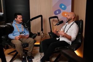 “Two men sit in leather chairs recording a podcast-style conversation. The man on the left wears a light blue hoodie, khaki pants, and an orange event lanyard, holding a notepad. The man on the right gestures while speaking into a microphone, wearing a white button-up shirt, black pants, and a black lanyard. A camera is visible in the foreground, and a banner with colorful speech bubble icons hangs behind them.