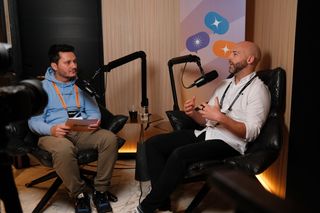 “Two men sit in leather chairs recording a podcast-style conversation. The man on the left wears a light blue hoodie, khaki pants, and an orange event lanyard, holding a notepad. The man on the right gestures while speaking into a microphone, wearing a white button-up shirt, black pants, and a black lanyard. A camera is visible in the foreground, and a banner with colorful speech bubble icons hangs behind them.