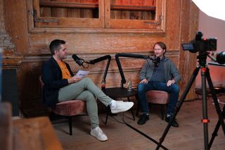 Two speakers recording a podcast-style interview during JoyConf, sitting across from each other in armchairs with microphones in a cozy wooden room.