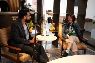 “Two people sit in armchairs recording a podcast-style conversation in a relaxed café setting. The man on the left has dark hair and a beard and is wearing glasses, a blazer, and a light blue shirt while speaking into a microphone. The woman on the right has short brown hair and is wearing a denim jacket, a green patterned shirt, and light pants, responding into another microphone. A small round table with cups of water sits between them, with shelves, plants, and a plush dog toy in the background.