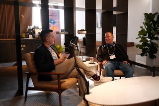Two men sit in armchairs recording a podcast-style conversation in a modern café setting. The man on the left wears a dark blazer, light pants, and an orange lanyard, holding a JoyConf notepad while speaking into a microphone. The man on the right wears a black long-sleeve shirt and jeans, listening and smiling with a Diet Coke can on the small table between them. Shelving, plants, and a coffee bar appear in the background.
