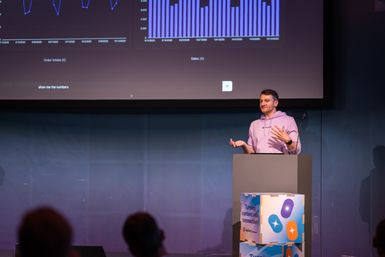 A person stands at a podium, gesturing while presenting data charts on a large screen in a dimly lit room.