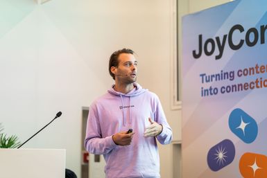 A person in a lavender hoodie speaks at a podium with a "JoyCon" banner in the background.