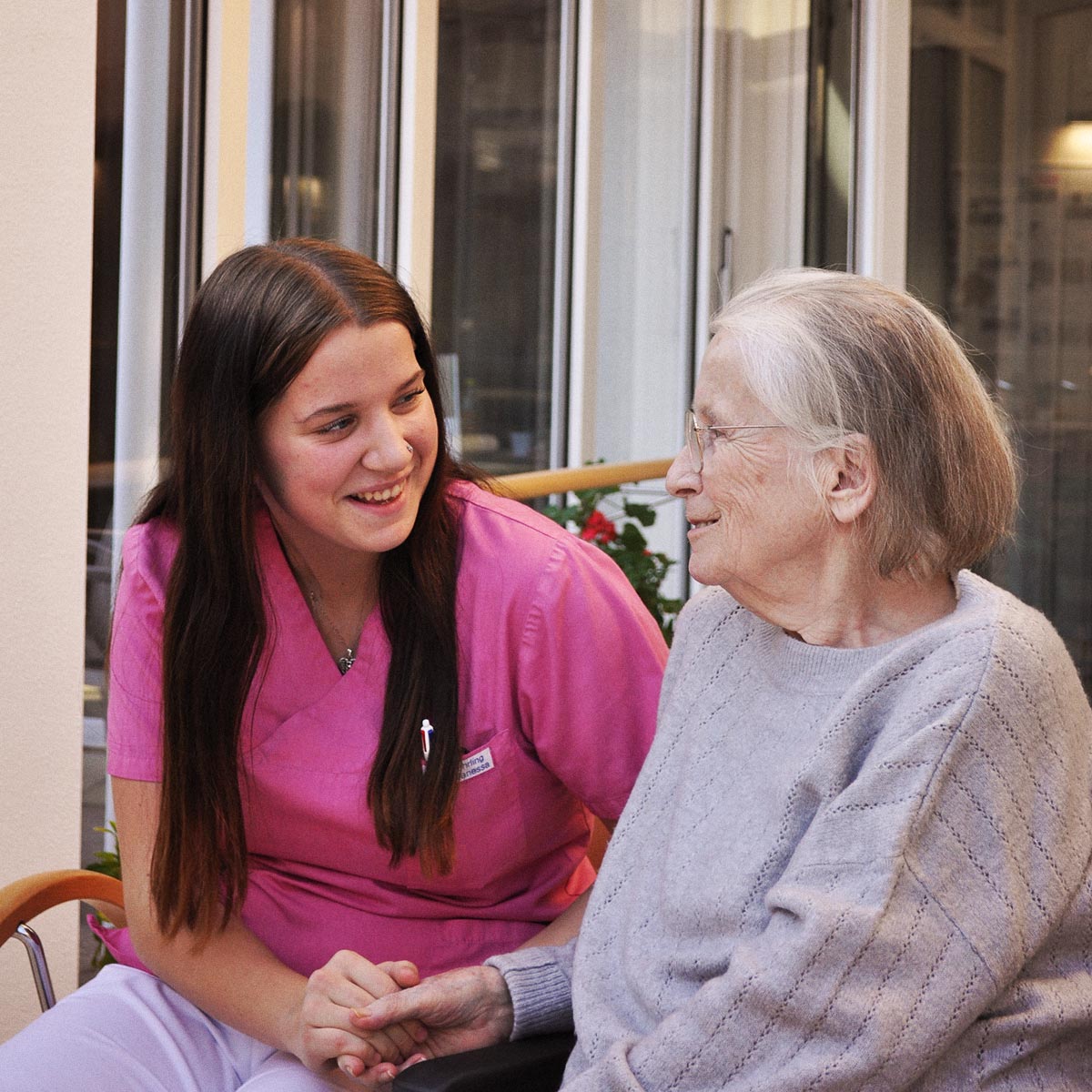 Eine junge Pflegekraft unterhält sich zugewandt mit einer Bewohnerin und hält ihre Hand.