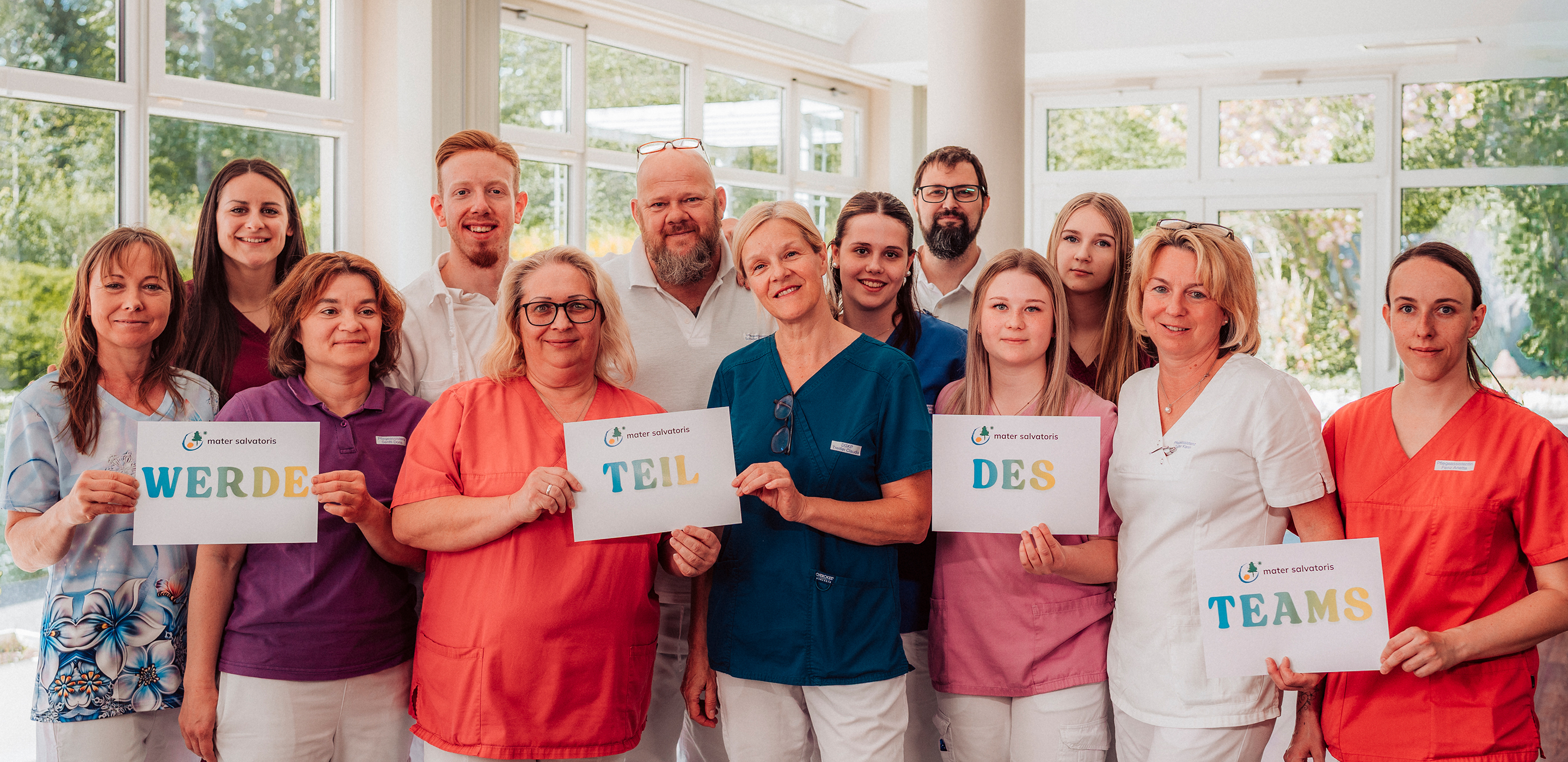 Ein diverses Team an Pflegekräften hält lächelnd Schilder mit Aufschrift "Werde Teil des Teams"