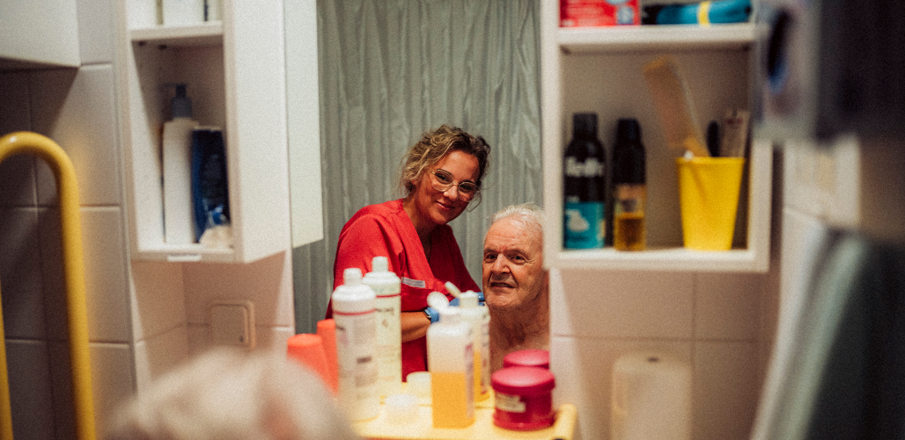Eine Pflegerin und ein Bewohner schauen sich im Badezimmer während der Körperpflege gemeinsam im Spiegel an