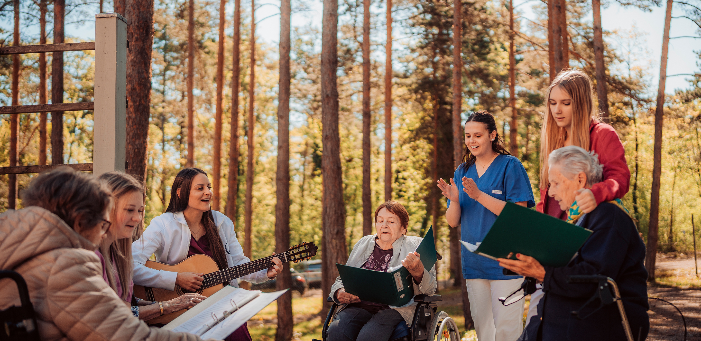 Eine Gruppe von jungen Pflegekräften und älteren Damen sitzen im Garten beisammen und singen mit Begleitung auf der Gitarre