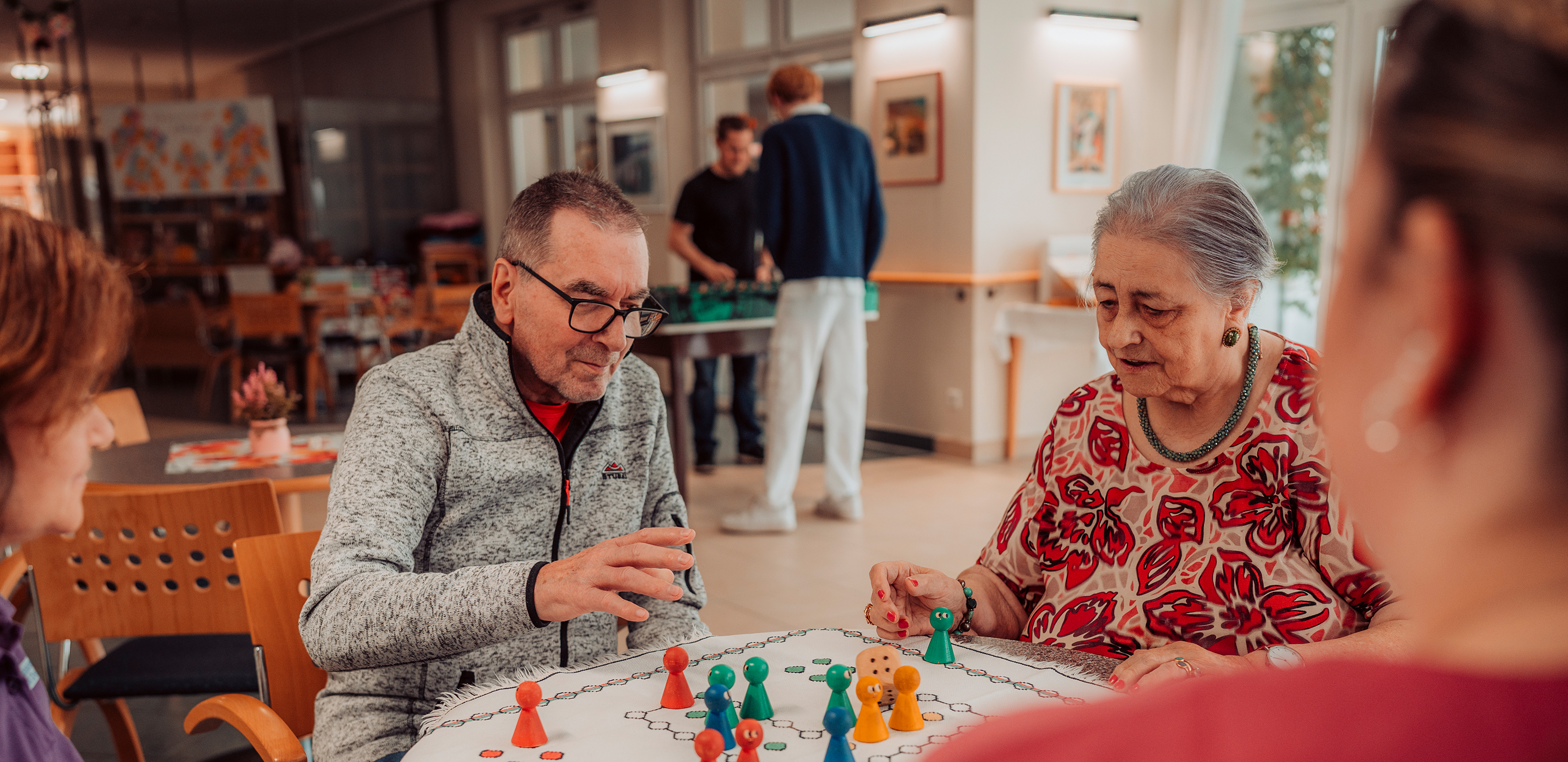 Zwei zufriedene ältere Menschen spielen mit zwei Betreuerinnen konzentriert ein Würfelspiel
