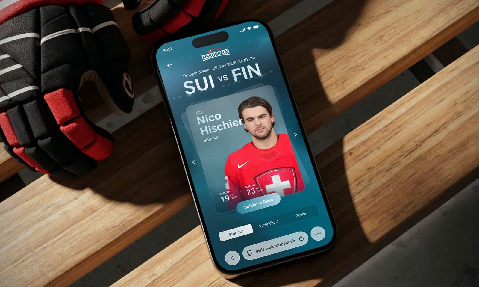In der Mitte liegt ein iPhone mit einem hellblauen Display. Auf dem Bildschirm ist die geöffnete Schweizer Website "swiss-eisrubbeln.ch" zu sehen. Angezeigt wird die Spielerauswahl eines Eishockey-Match der Gruppenphase am 26. Mai 2026: SUI vs. FIN (Schweiz gegen Finnland).