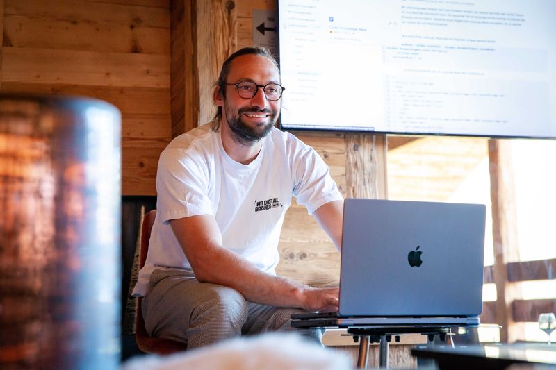 Mann mit Brille sitzt vor einem MacBook mit Präsentation im Hintergrund.