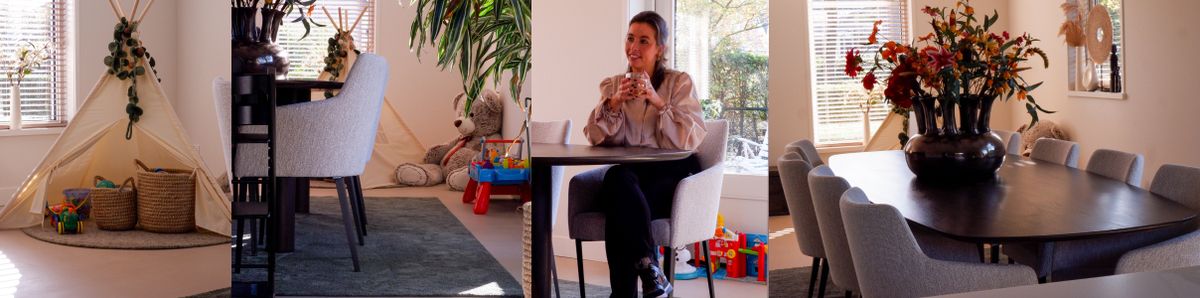 Een vrouw zit aan een tafel in een modern ingerichte kamer met een tipi en speelgoed in de hoek, en een grote vaas bloemen op de achtergrond.