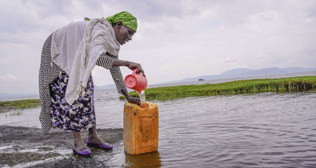 Vrouw met groene hoofddoek giet water uit een rode kan in een oranje jerrycan bij een meer met bergen op de achtergrond.