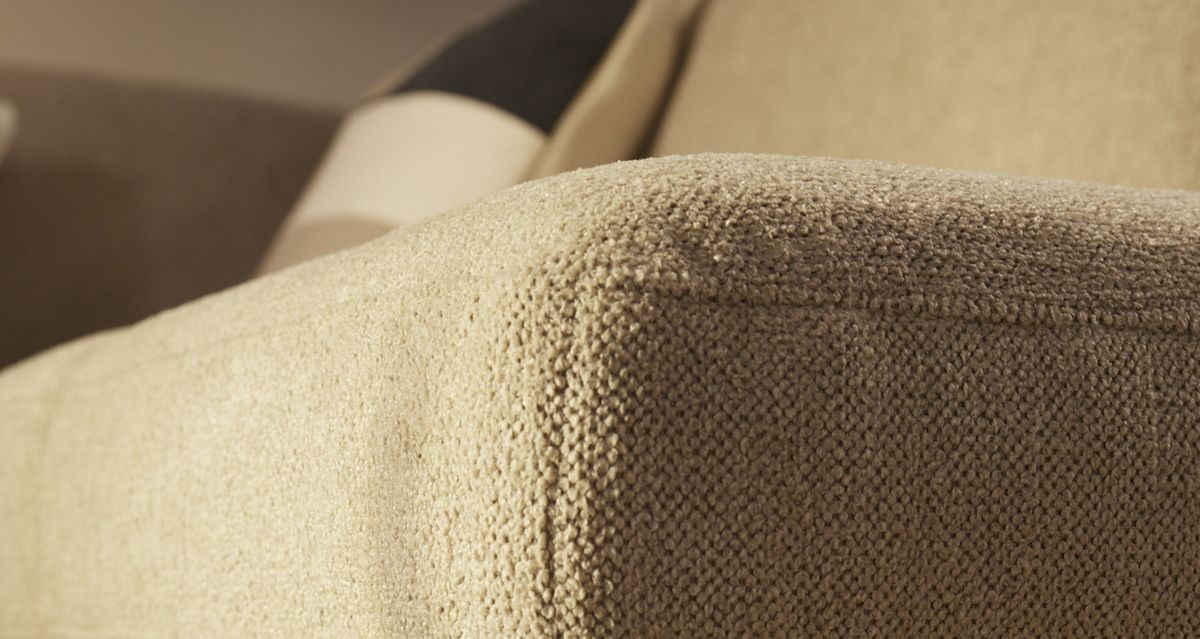 Beige, stoffen hoek van een bank in close-up, met een kussen op de achtergrond.