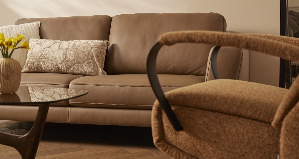 Bruin lederen bank met kussen, houten salontafel met gele bloemen in vaas en een comfortabele stoel op een houten vloer.