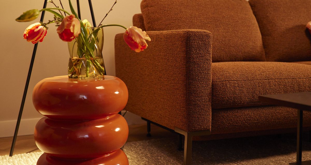 Bruin stoffen bank, ronde oranje bijzettafel met vaas en oranje tulpen in een sfeervol verlichte woonkamer.