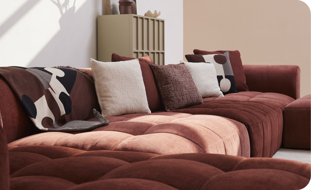 Roze-rode, fluwelen loungebank met diverse kussens in neutrale tinten en geometrische patronen in een moderne, minimalistische kamer.