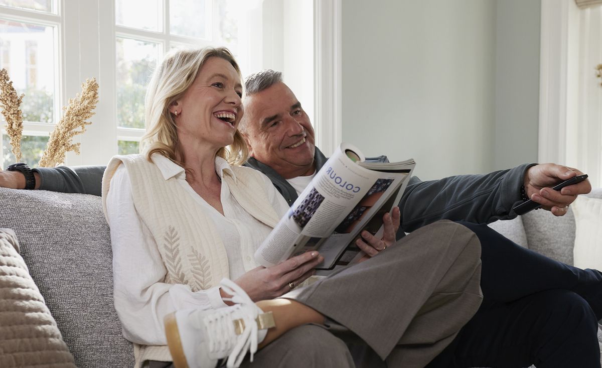 Een lachend koppel zit op de bank, de vrouw leest een tijdschrift terwijl de man de afstandsbediening vasthoudt.