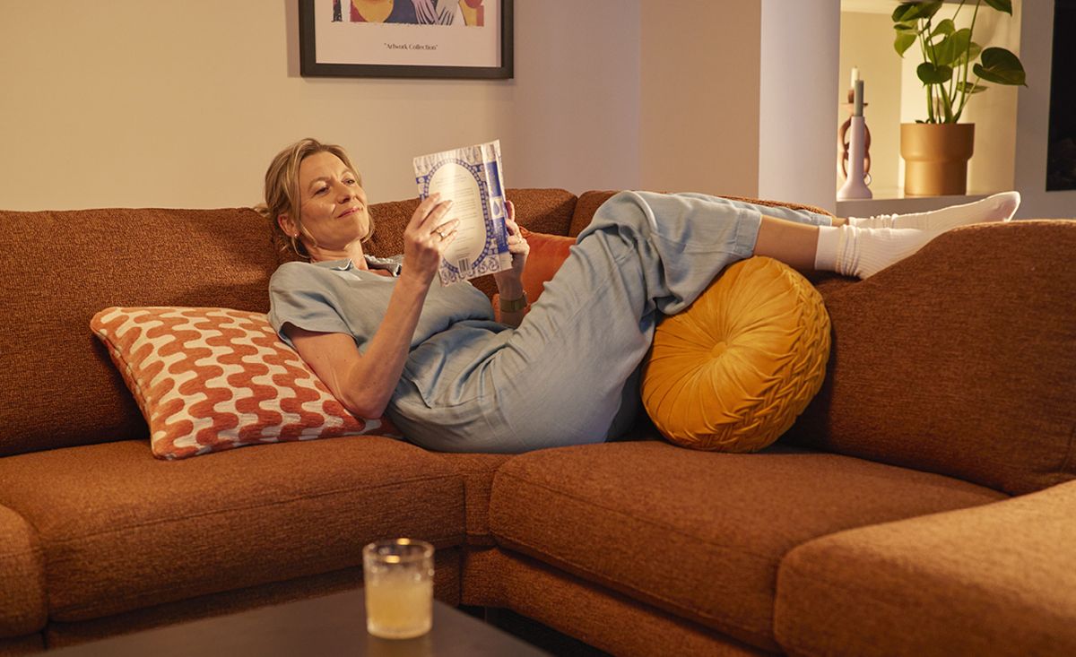 Vrouw in blauwe outfit ligt ontspannen op een oranje bank, leest een boek en heeft kussens om zich heen. Een drankje staat op de tafel.