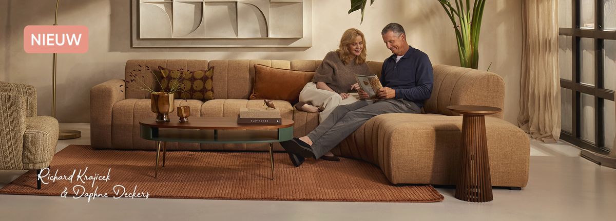 A couple sits on a beige sectional sofa reading a magazine, in a cozy living room with warm decor and a wooden coffee table.
