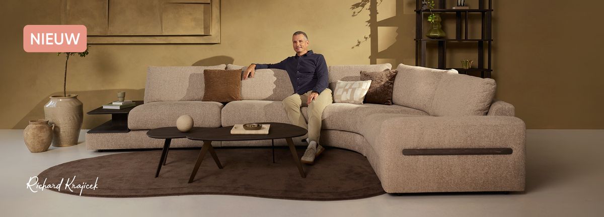 A man in a navy shirt and beige pants sits on a large beige sectional sofa in a modern living room with decorative elements.