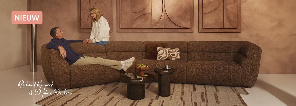 A man reclines on a brown sofa while a woman leans over him, in a warm, art-filled living room with a modern design.
