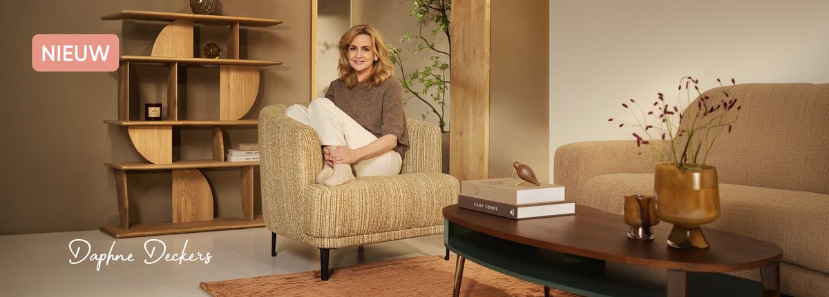 A woman sits on a beige armchair in a stylish living room with modern decor, including a wooden shelf and a table with books and a vase.