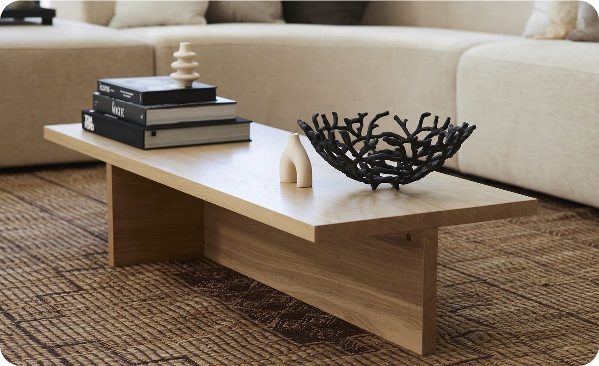 Houten salontafel met stapel boeken, abstracte sculptuur en zwart geometrisch metalen mand, op een tapijt in een woonkamer.