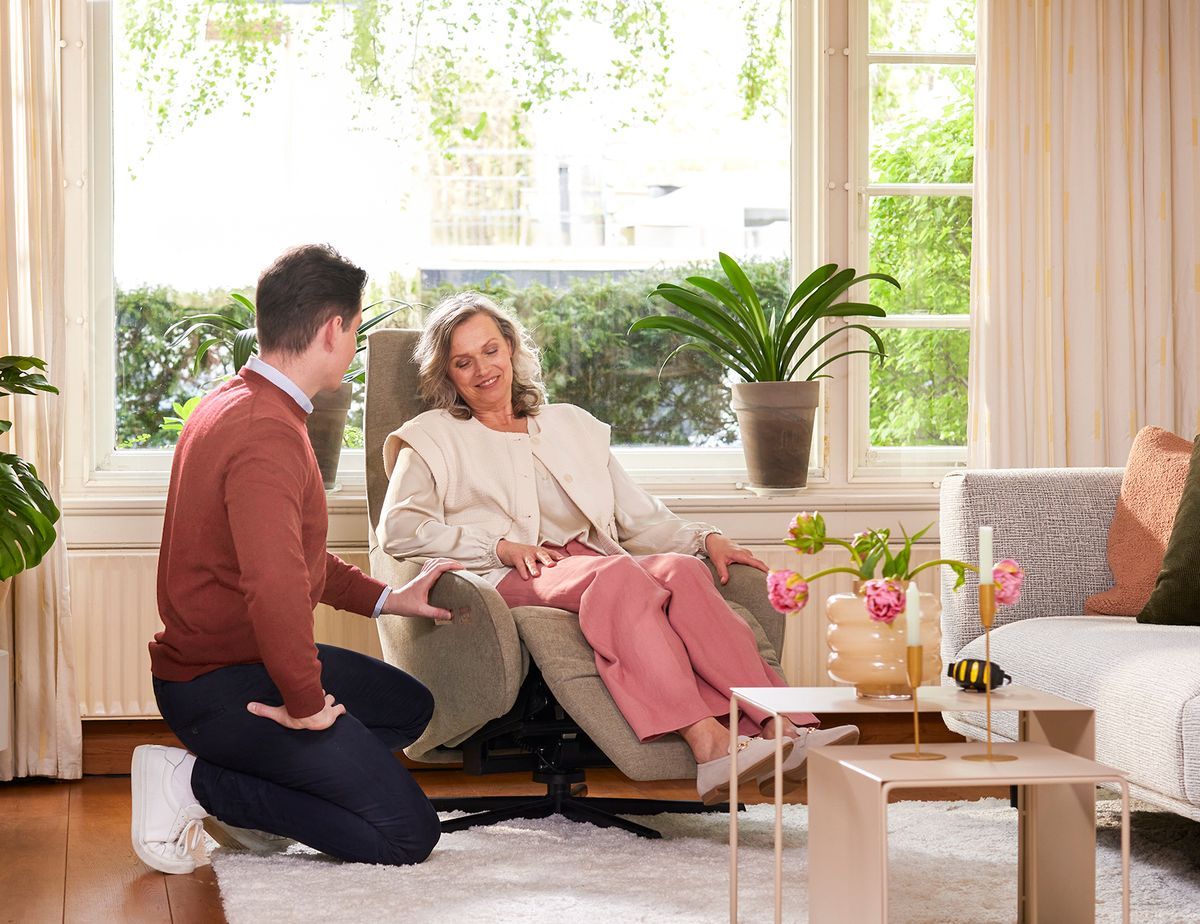 A woman relaxes in a chair, smiling, while a man kneels beside her in a cozy living room with plants and a large window.