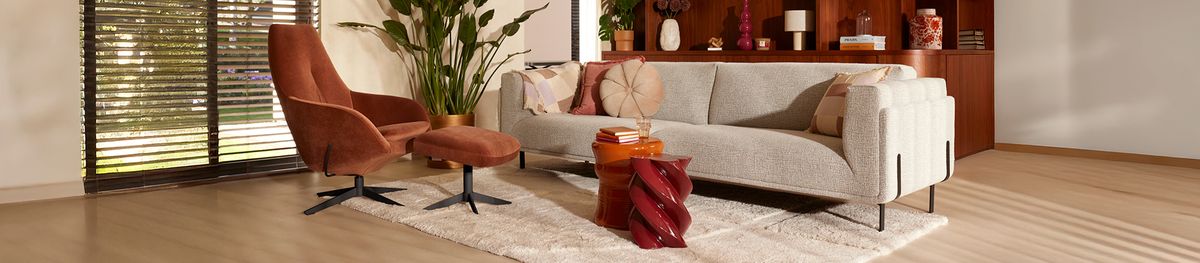 Moderne woonkamer met een lichtgrijze bank, bruine fauteuil en poef, tapijt, planten en houten kast. Houten vloer en grote ramen met jaloezieën.