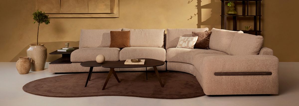 Modern living room with a beige sectional sofa, earthy-toned cushions, dark wood coffee table, and decorative vases on a brown rug.
